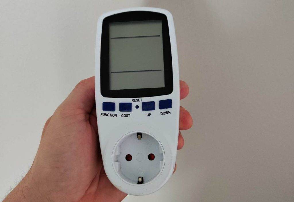 Energiemessgeräte-Test: Hand hält ein weißes Modell mit vier schwarzen Funktionstasten und einer Stecker-Buchse ausgeschaltet vor einer hellen Wand.