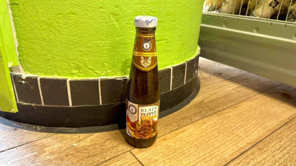 Eine Flasche Pfeffersauce steht zum testen vor einer grünen Wand mit schwarzem Sockel.