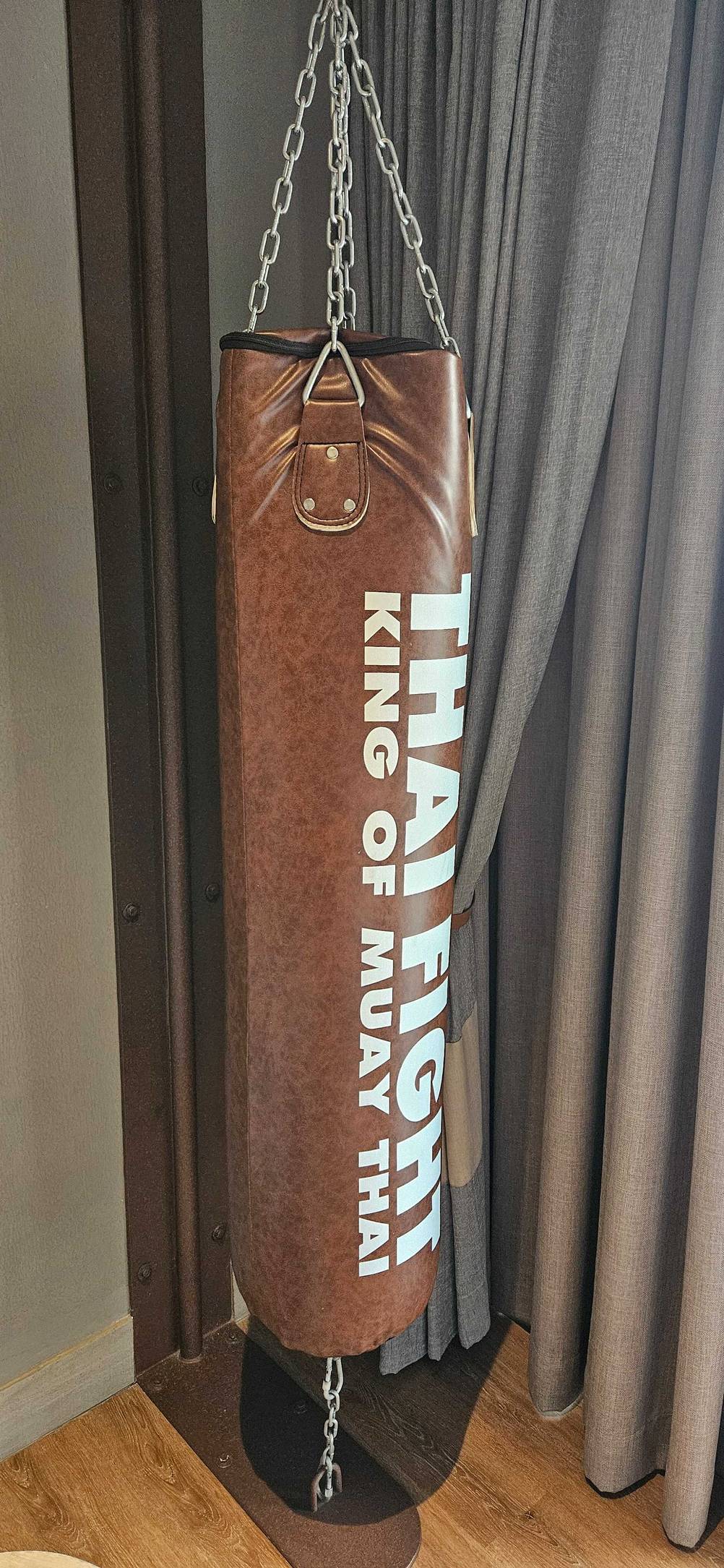 Ein getesteter Boxsack mit Befestigung vor einem Vorhang auf einem Holzboden.