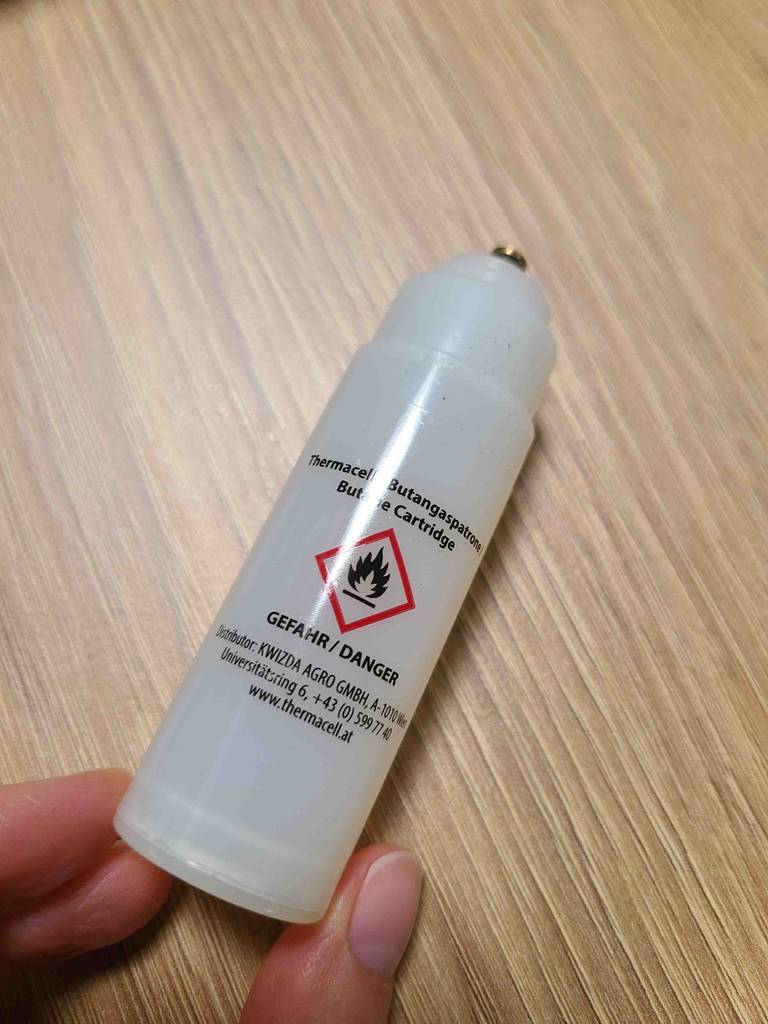 Thermacell-Mückenabwehr im Test: eine kleine Gaskartusche inklusive Anschluss liegt zwischen Daumen und Zeigefinger auf einer Holzoberfläche.