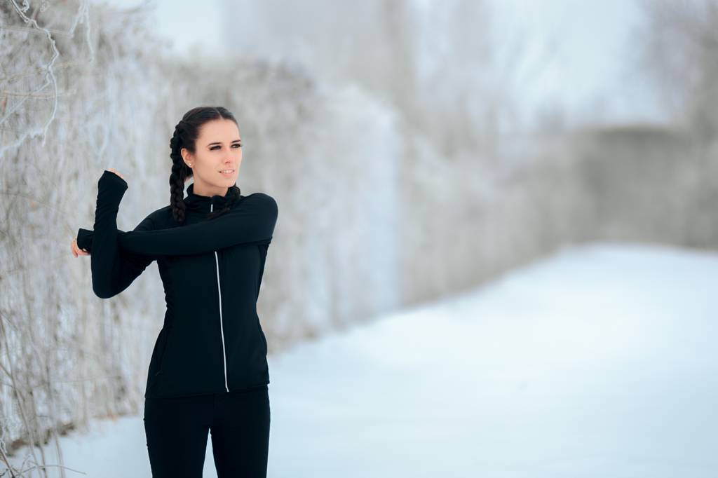 Thermo-Laufhose-Damen-Test: Eine Frau in schwarzer Laufkleidung dehnt sich in einer winterlichen Landschaft. 
