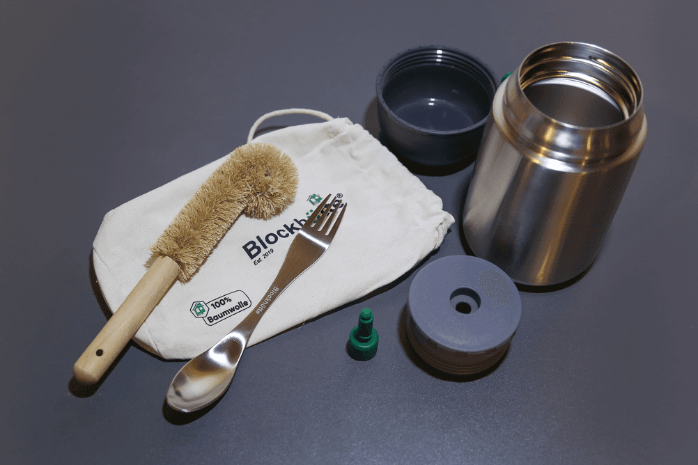 Test der Thermo-Lunchboxen: Neben der Thermo-Lunchbox von Blockhütte liegen der Verschluss, der Deckel, das Ventil, eine Löffel-Gabel-Kombi, eine Reinigungsbürste und ein Transporttäschchen.