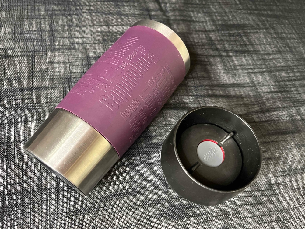 Ein getesteter Thermobecher mit seinem Deckel daneben auf einer grauen Tischdecke.