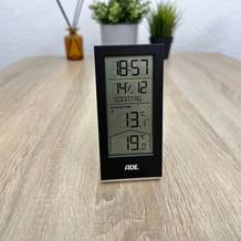 Ein Ade-WS2324-Thermometer steht aufrecht auf einem Tisch und zeigt Uhrzeit, Datum sowie Innen- und Außentemperatur auf dem Display an.