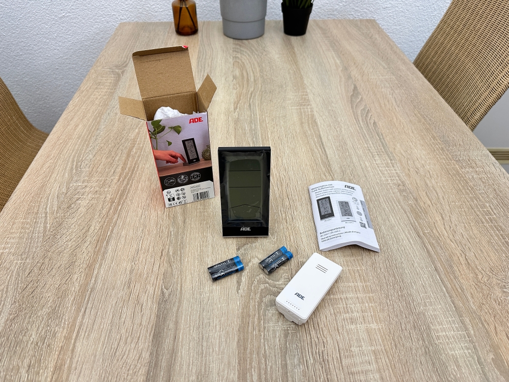 Der Lieferumfang eines Ade-WS2324-Thermometers liegt ausgepackt auf einem Holztisch mit Hauptgerät, Außensensor, Batterien, Anleitung und Verpackung.
