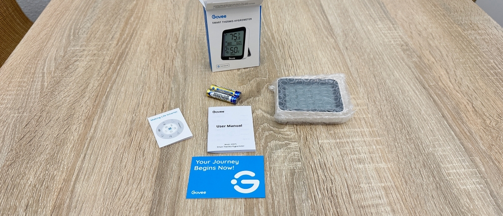 Der Lieferumfang eines Govee-H5075001DE-Thermometers liegt vollständig ausgepackt auf einem Tisch mit Gerät, Batterien, Anleitung, Zubehör und Verpackung.