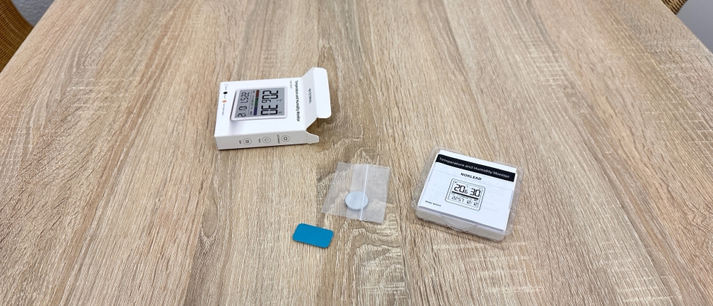 Der Lieferumfang eines Noklead-5253M-FR-Thermometers liegt ausgepackt auf einem Tisch mit Gerät, Verpackung, Klebepad und Zubehör.