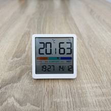 Ein Noklead-5253M-FR-Thermometer steht frontal auf einem Holztisch und zeigt Temperatur, Luftfeuchtigkeit, Komfortanzeige sowie Uhrzeit auf dem Display an.