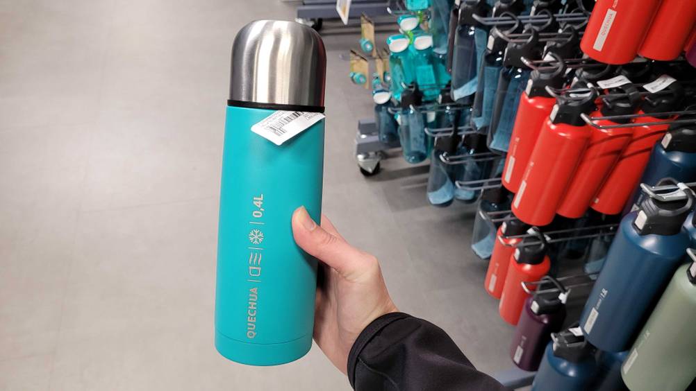Eine Hand hält eine getestete Thermosflasche vor ein Regal mit vielen anderen.