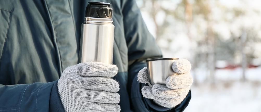 Eine Person hält mit Handschuhen eine Thermosflasche mit Becher in der Hand.