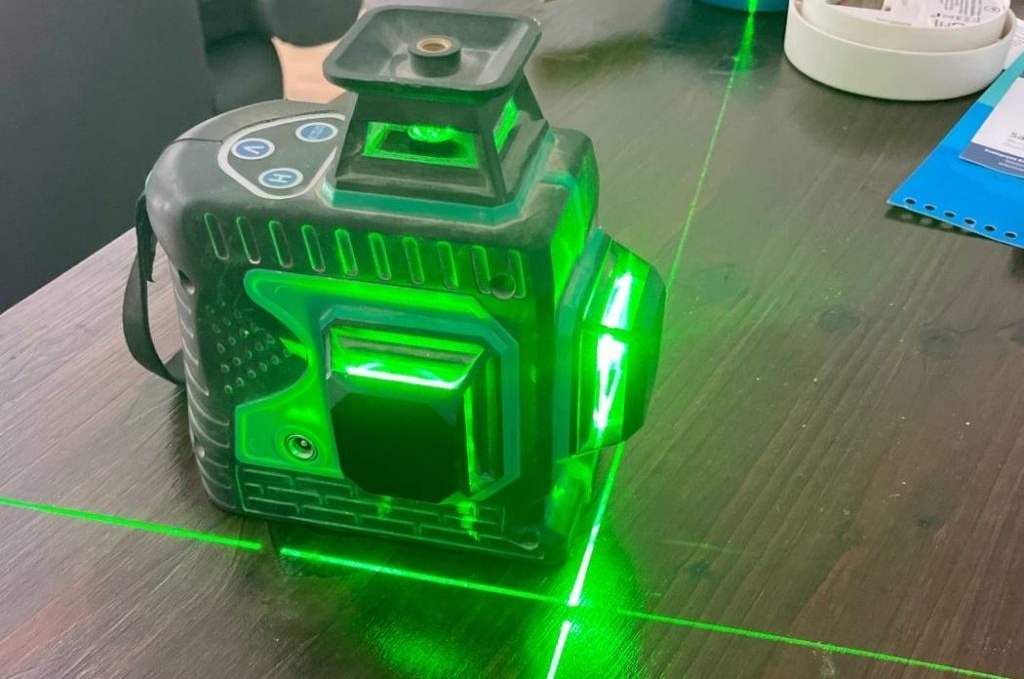 Batteriebetriebener, getesteter Kreuzlinienlaser in Funktion mit grünem Laserlicht steht auf einem Holztisch.