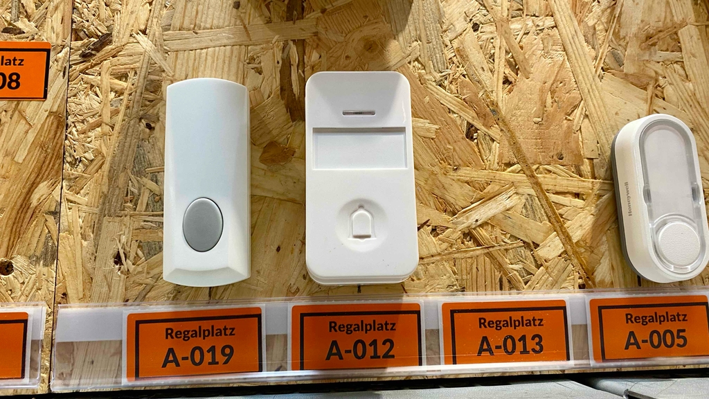 Mehrere weiße Funkklingeln im Test sind auf einer Holzwand im Geschäft über ihren orangen Produktcode-Etiketten ausgestellt.