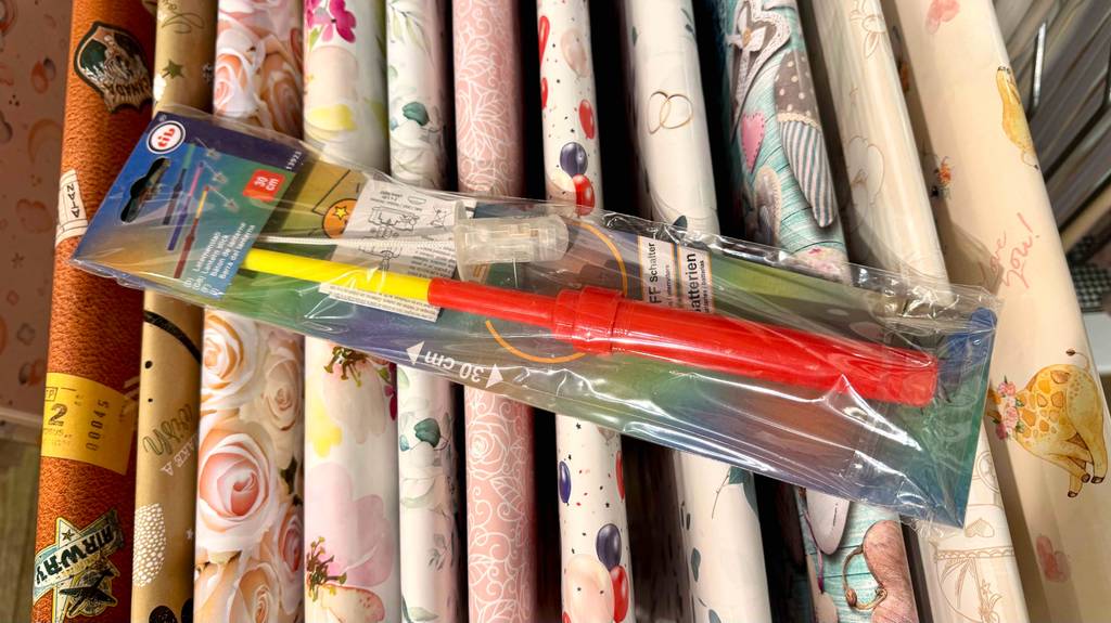 Blick auf den Griff von oben eines in Plastik verpackten Laternstabes, der zum testen auf Geschenkpapierrollen liegt.