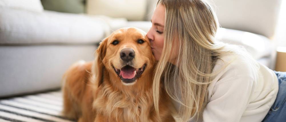 Junge Frau und ein Golden Retriever liegen auf einem Teppich und die Frau küsst den Hund auf die Wange.