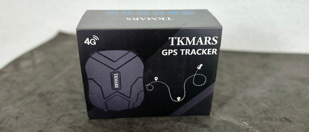 Die Verpackung des TKMARS GPS Trackers steht aufrecht auf einem grauen Untergrund. Aufdrucke zeigen das Logo und technische Icons.