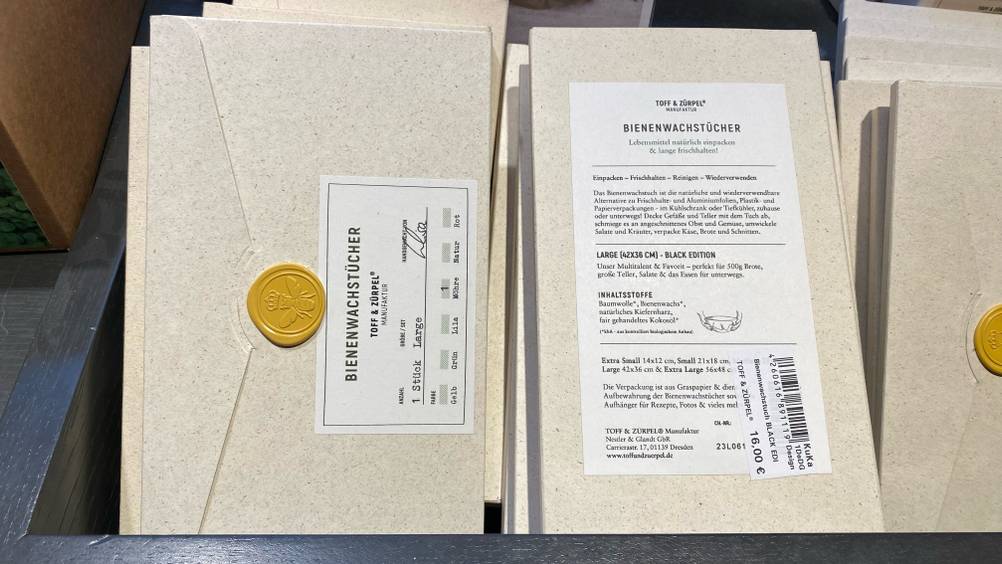 Bienenwachstuch im Test: mehrere Produkte in hellen Umschlägen verpackt, befinden sich mit Fokus auf die Vorderseite inklusive gelbem Bienen-Siegel und auf die Rückseite mit Produktbeschreibung in einem Verkaufsregal.