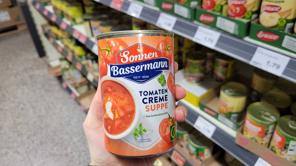 Eine Hand hält eine Dose der getesteten Tomatensuppe-Dose vor ein Regal.