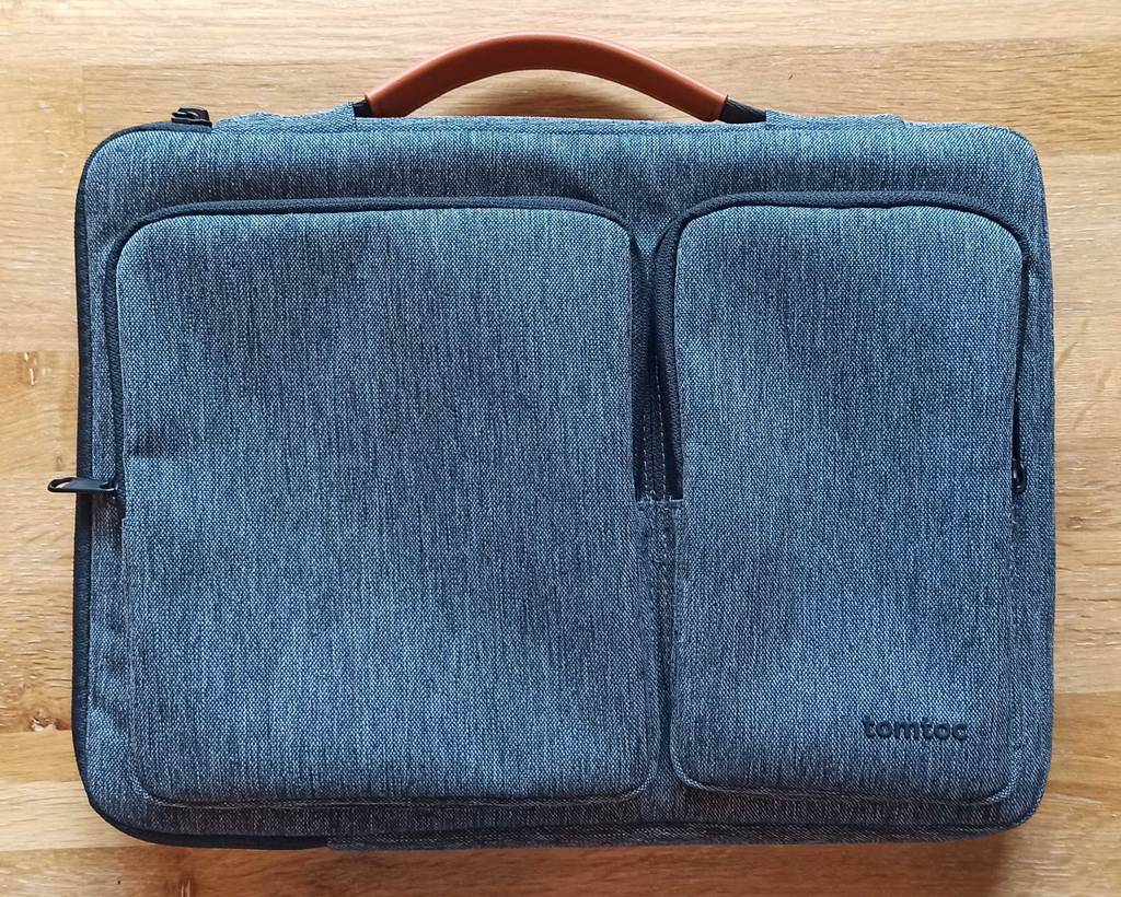 Grau-blau melierte getestete MacBook-Tasche von oben inklusive zwei Außentaschen und Griff in Braunleder-Optik liegt auf einem Holzuntergrund.