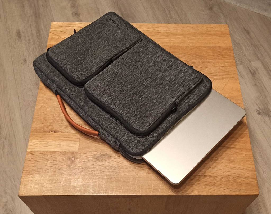Grau melierte getestete MacBook-Tasche inklusive halb eingestecktem silberfarbenem MacBook liegt auf einem Holzhocker.