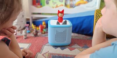 Die hellblaue Toniebox 2 mit einer roten Toniefigur steht auf einem Teppich im Kinderzimmer.
