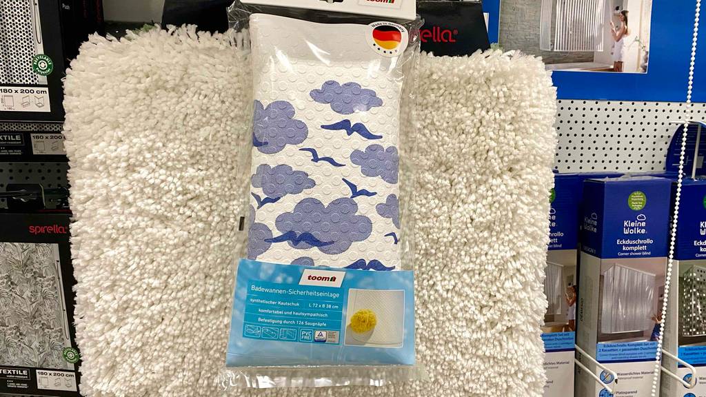 Badewannenmatten-Test: ein Toom Modell in Weiß mit blauen, stilisierten Wolken und Vögeln hängt in seiner transparenten Verpackung über einem weißen Badezimmerteppich im Verkaufsbereich.