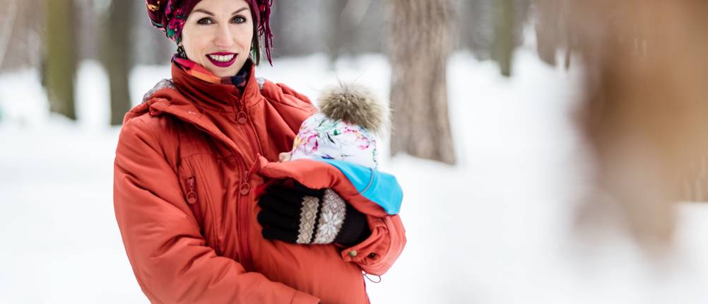tragejacke-herren-test: Eine Frau trägt ein Baby mit weißer Pudelmütze in einer Tragejacke.