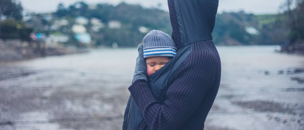 tragejacke-herren-test: Person steht vor Flußlandschaft und trägt das Baby unter der Jacke.