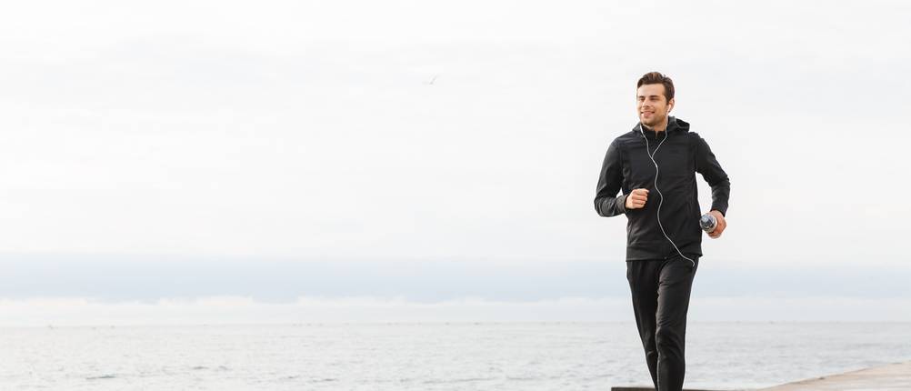 Trainingsanzug im Test: Junger Mann im schwarzen Trainingsanzug joggt einen Strand entlang.