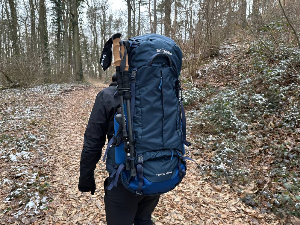 Eine Person trägt den Tatonka Yukon 60+10 mit befestigten Trekkingstöcken auf einem Waldweg. Der Rucksack ist vollständig beladen.