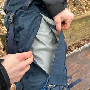 Eine Hand öffnet das seitliche Frontfach des Tatonka Yukon 60+10. Die graue Innenverkleidung des Hauptfachs wird sichtbar.