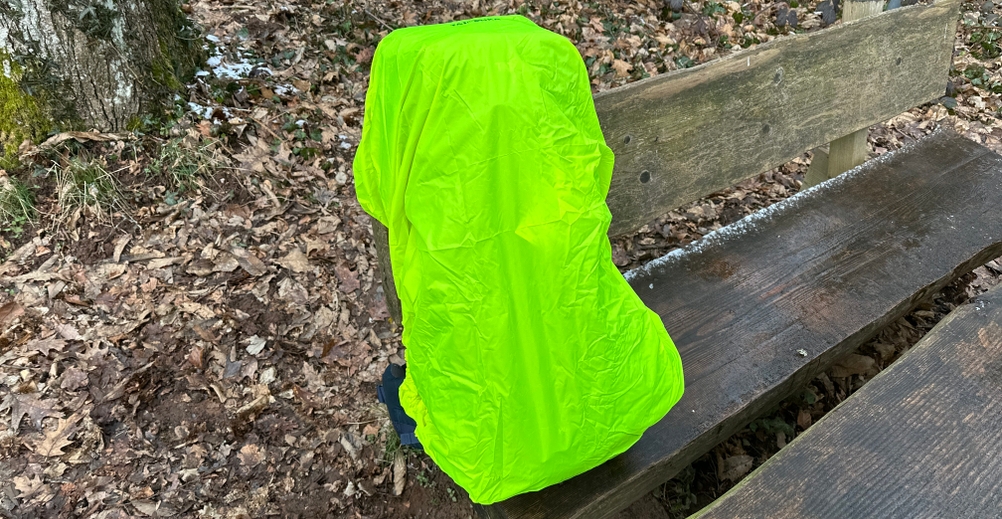 Der Tatonka Yukon 60+10 ist mit einer leuchtend gelben Regenhülle überzogen. Der Rucksack steht auf einer Bank im Wald.