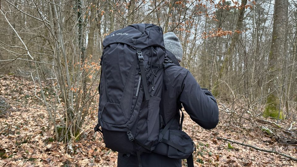 Der Vaude Asymmetric 42+8 wird von einer Person im Wald getragen. Der Rucksack liegt eng am Rücken an.