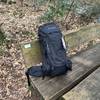 Der Vaude Asymmetric 42+8 steht vollständig gepackt auf einer Holzbank im Wald. Der Trekkingrucksack ist von vorn zu sehen, inklusive Etikett.