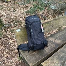 Der Vaude Asymmetric 42+8 steht vollständig gepackt auf einer Holzbank im Wald. Der Trekkingrucksack ist von vorn zu sehen, inklusive Etikett.