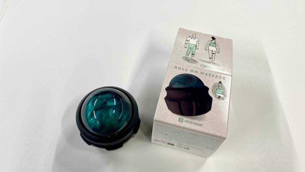 Ansicht von oben auf den grünen Roller eines getesteten Handmassagegeräts, das neben seiner Originalverpackung auf einer weißen Fläche liegt.