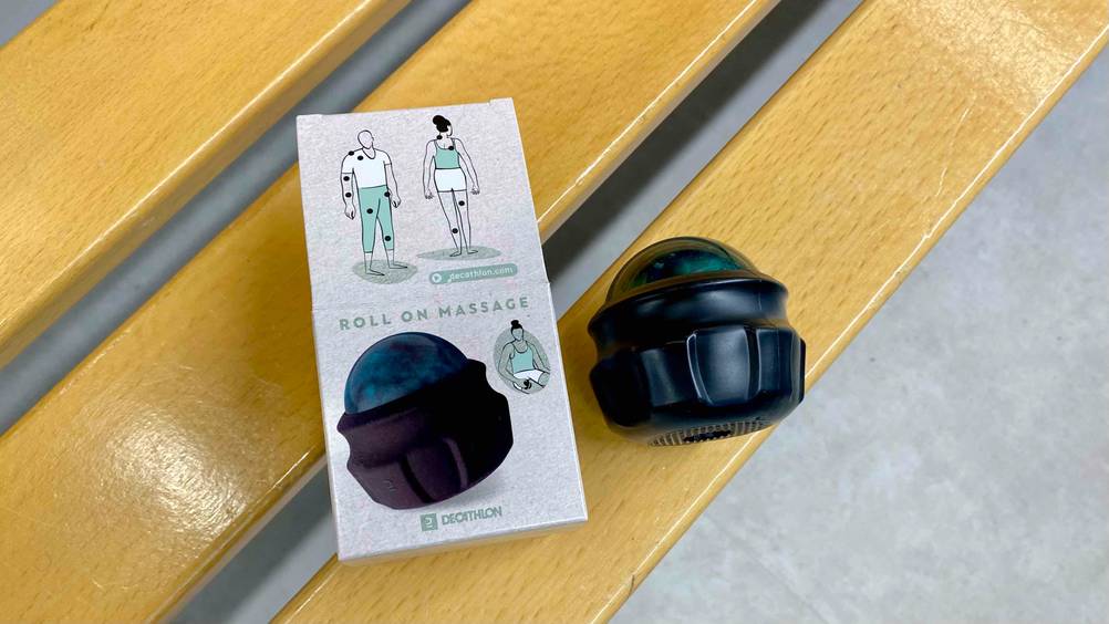 Im Test: ein Handmassagegerät in Schwarz mit grünem Roller liegt auf der Seite neben seiner Originalverpackung auf einer Holzbank.