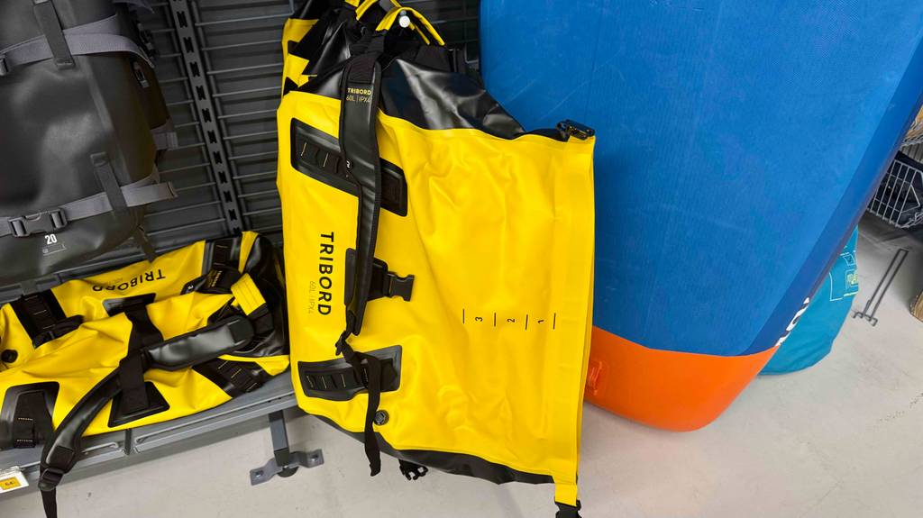 Getestet: Die wasserdichte Reisetasche in Gelb mit 60 Liter Füllvolumen lehnt an einem blauen Surfbrett im Sportwarengeschäft.