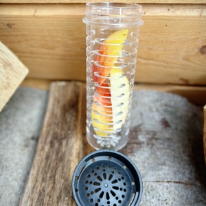 Der Fruchteinsatz der 720°Dgree uberBottle liegt neben dem Siebdeckel auf einem Holzuntergrund. In der transparenten Kartusche befinden sich Zitronen- und Apfelscheiben.