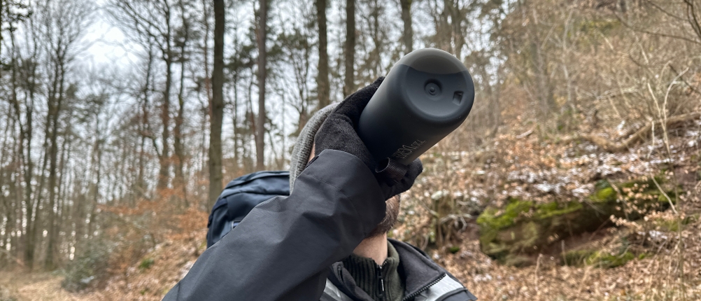 Ein Wanderer trinkt aus der 720°Dgree-uberBottle-Trinkflasche im Wald. Die schwarze Flasche wird mit Handschuhen sicher gehalten.
