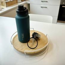 Eine Trinkflasche mit Zubehör steht auf einer runden Scheide auf enem weißen Tisch.