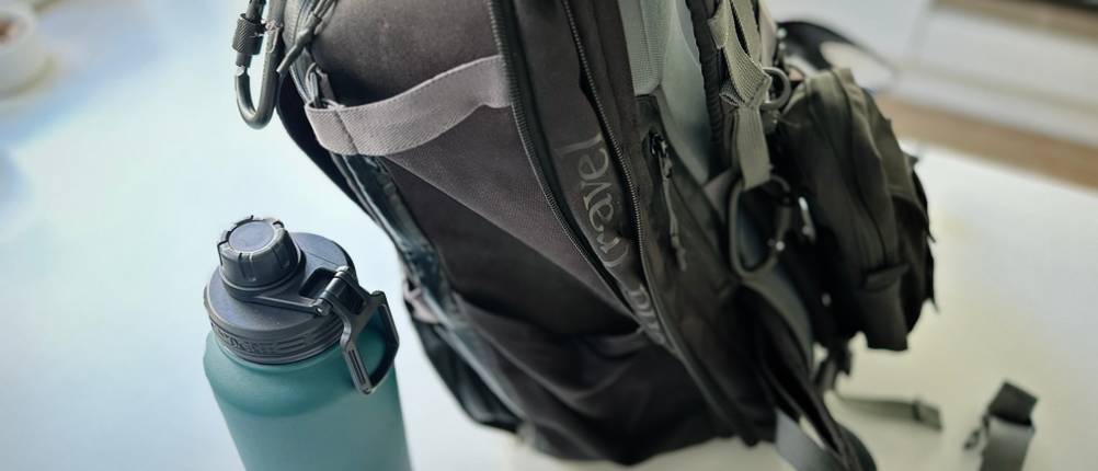 Die Trinkflasche steht auf einem weißen Untergrund neben einem Rucksack.