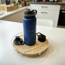 Eine Trinkflasche mit Zubehör steht auf einer runden Scheide auf enem weißen Tisch.