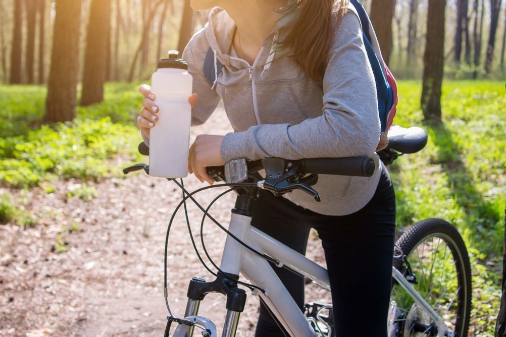 Trinkflasche-für-kohlensäurehaltige-Getränke: eine Frau steht mit ihrem Fahrrad im Wald und hält eine Trinkflasche in der Hand.