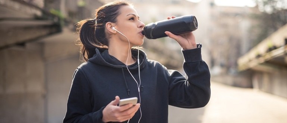 Trinkflasche-für-kohlensäurehaltige-Getränke-Test: Frau steht mit einem Handy in der Hand und trinkt aus einer Flasche