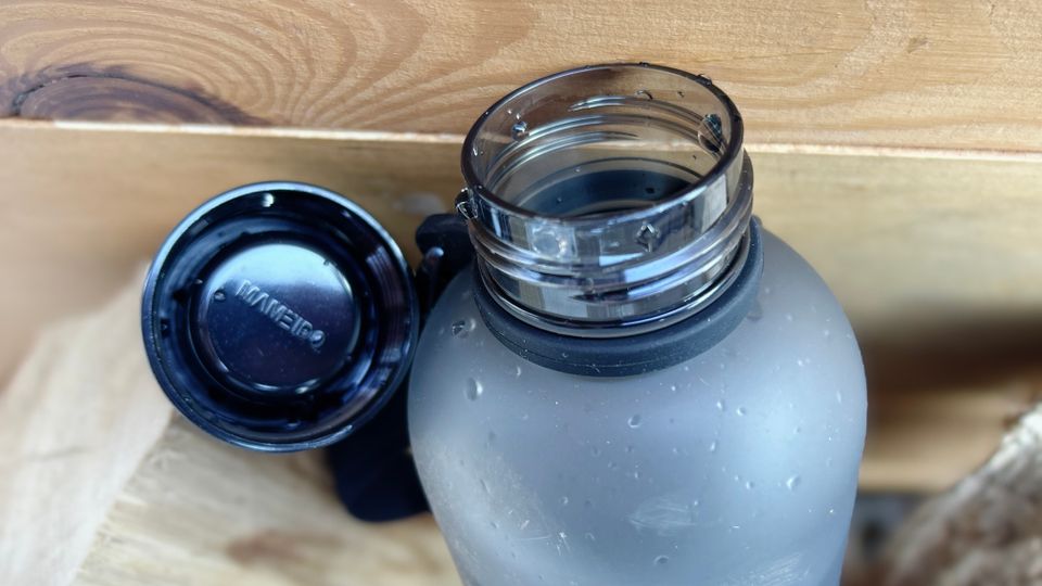 Die Mameido Tritan Trinkflasche steht geöffnet neben ihrem Deckel. Der Schraubverschluss liegt daneben.