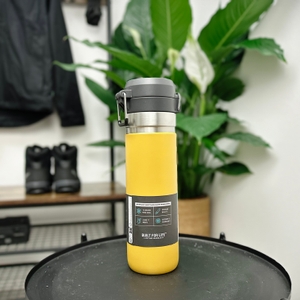 Die Rückseite der Stanley Quick Flip Flasche zeigt Angaben zur Isolierleistung und Garantie. Die Flasche steht auf einem Tisch vor einer Pflanze.