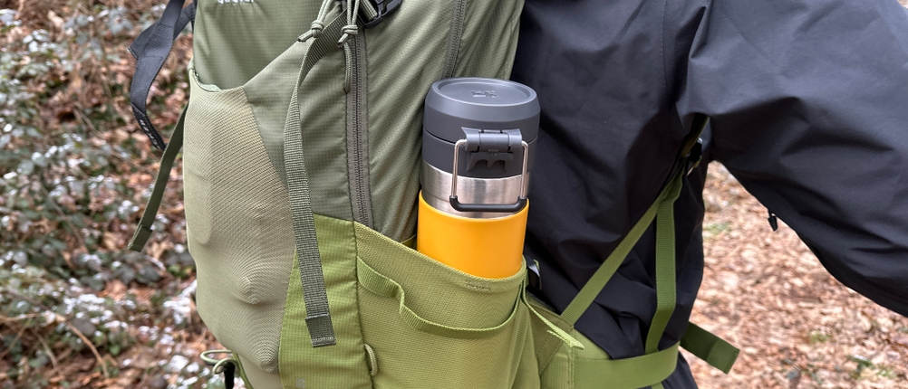 Die Stanley-Quick-Flip-Trinkflasche steckt in der Seitentasche eines grünen Rucksacks. Eine Person trägt den Rucksack im Wald.