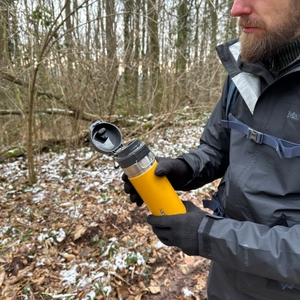 Eine Person hält die Stanley-Quick-Flip-Flasche mit geöffnetem Deckel in einem Waldgebiet. Der Mechanismus ist sichtbar.