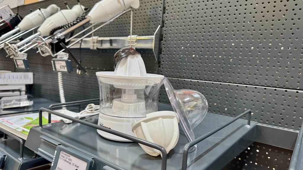 Die weiße getestete elektrische Zitruspresse steht inklusive Zubehör auf einem grauen Ausstellungsregal.