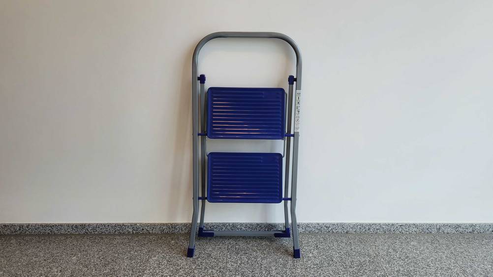 Eine zusammengeklappte 2-Stufen-Trittleiter im Test lehnt an einer hellen Wand.